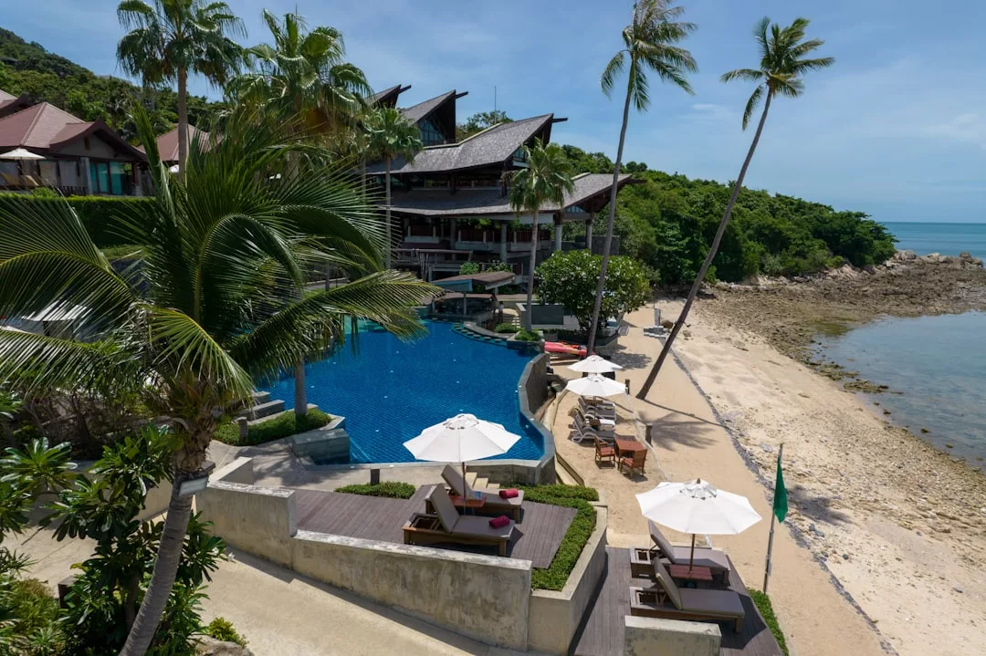 A beachfront real estate resort with a swimming pool, sun loungers under umbrellas, palm trees, and a sandy beach beside clear, calm sea water.