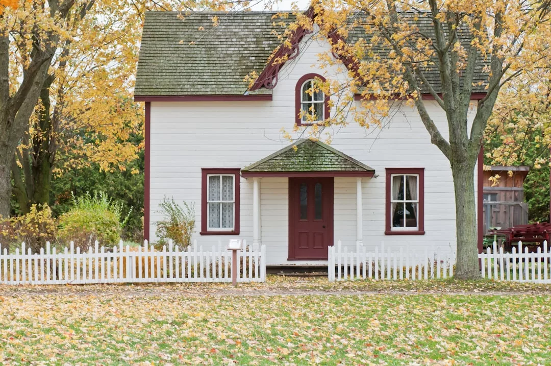 A charming white two-story real estate gem with red trim, a covered front porch, and a white picket fence, nestled among autumn trees with golden yellow leaves.