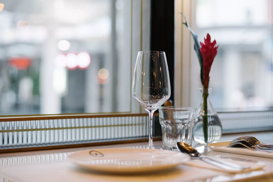A table set with a wine glass, water glass, plate, cutlery, and a small vase with a red flower sits beside a window—perfect for showcasing elegant dining in real estate photography.