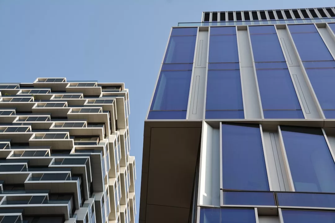 Two modern high-rise buildings with glass facades and geometric designs rise impressively against a clear blue sky, showcasing striking real estate architecture.
