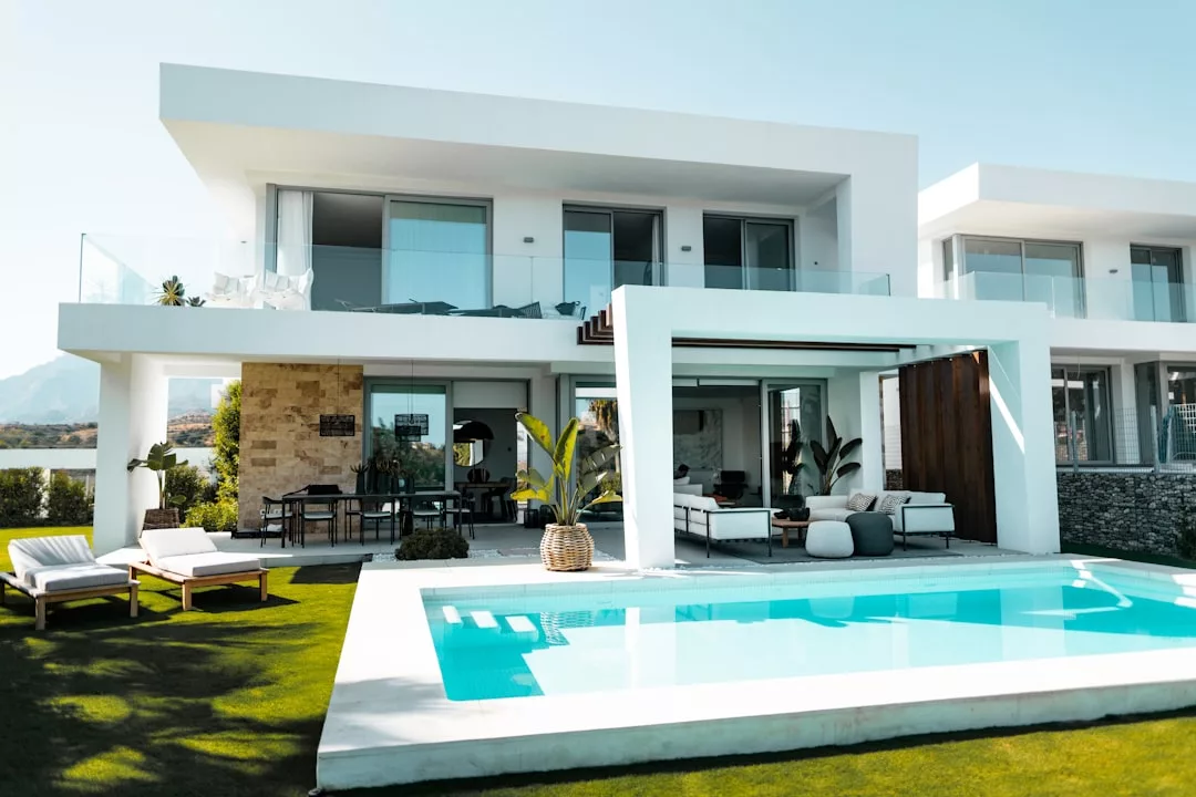 Modern two-story real estate with a swimming pool, sun loungers, outdoor seating area, large windows, and a well-kept lawn under clear skies.
