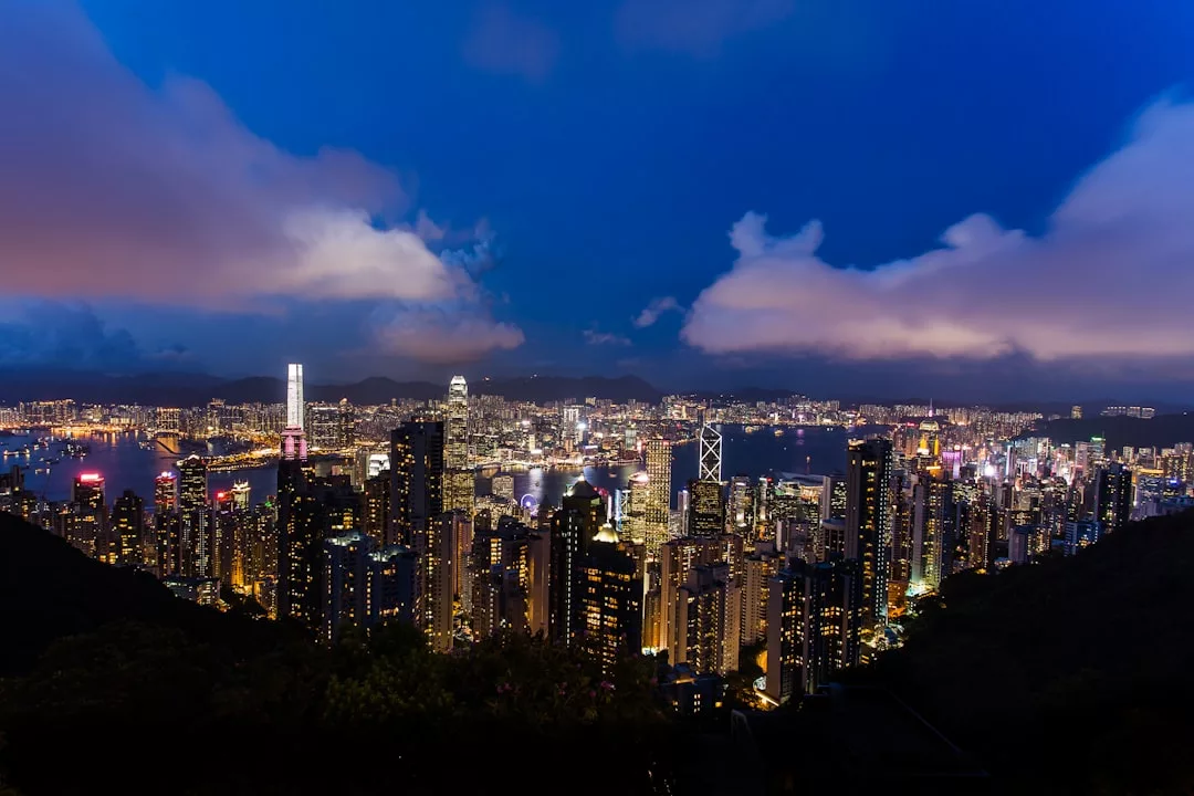 A nighttime cityscape shows a dense cluster of illuminated skyscrapers and buildings under a blue sky with clouds, highlighting the vibrant energy of urban real estate.