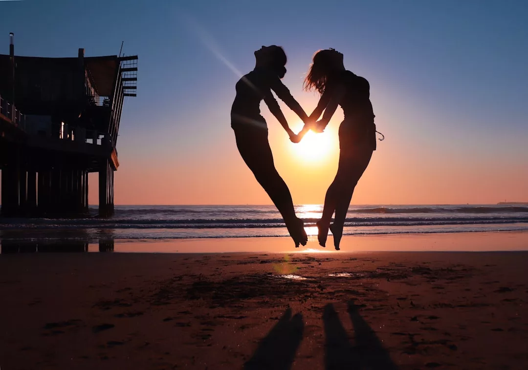 Two people jump on a beach at sunset, holding hands and forming a heart shape with their bodies in front of the sun—a perfect moment that captures the joy of finding your dream real estate by the sea.