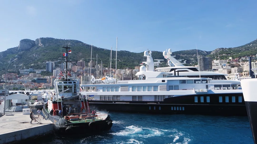 Two boats are docked at a marina in front of hillside city real estate, with mountains and a clear blue sky in the background.