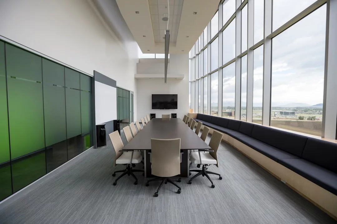 Modern conference room in prime real estate with a long table, twelve chairs, large windows on one side, a wall-mounted TV, and a striking green glass wall opposite.