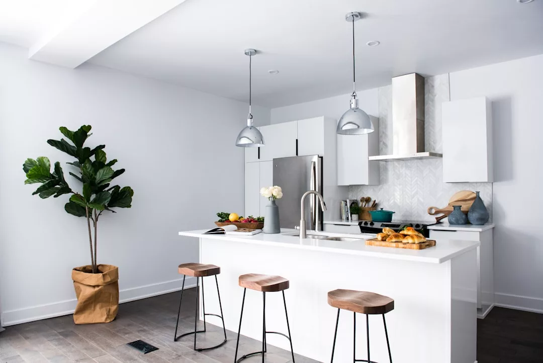 Modern kitchen with a white island, three wooden stools, stainless steel appliances, pendant lights, potted plant, and various kitchen items—perfect for showcasing your real estate listing.