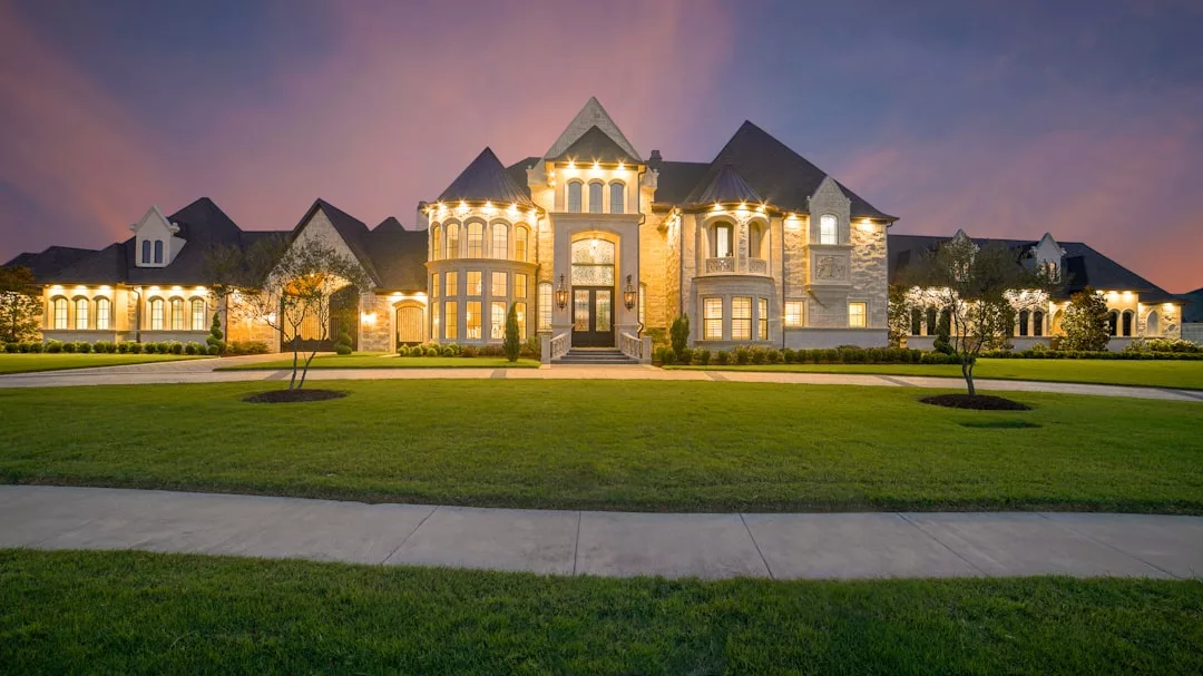 Large, illuminated real estate mansion with extensive front lawn and multiple peaked roofs at dusk, seen from across a wide driveway.