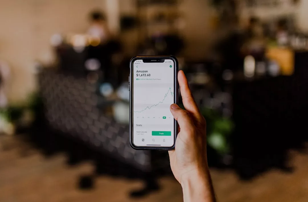 A hand holds a smartphone displaying a stock trading app with an upward trending chart for Amazon. The blurred background hints at real estate elements, connecting digital finance with property investment.