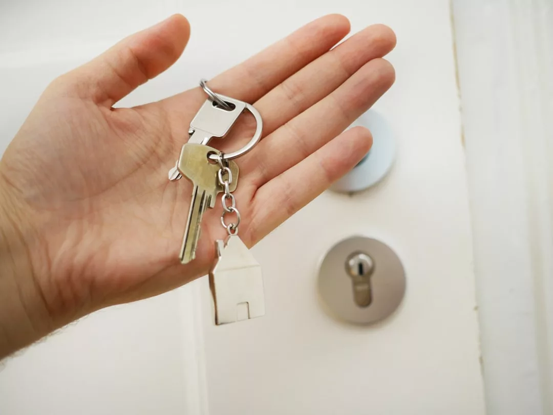 A hand holds two silver keys and a house-shaped keychain, symbolizing a fresh start in real estate, in front of a white door with a silver lock.