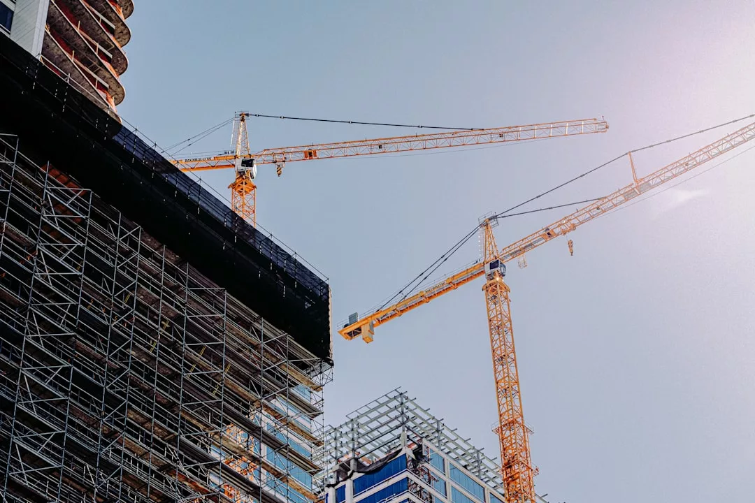 Two tall yellow cranes stand beside a partially constructed real estate building covered in scaffolding under a clear sky.