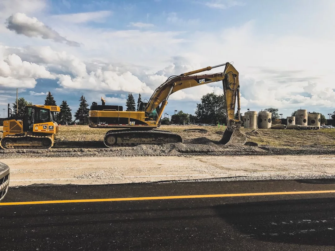A yellow excavator and a bulldozer work on a real estate construction site near a road, with concrete pipes and trees in the background under a partly cloudy sky.
