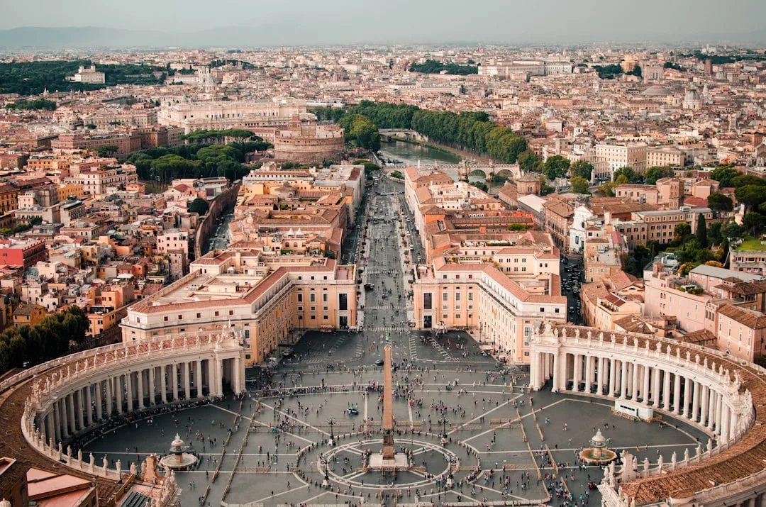 Aerial view of St. Peter’s Square in Vatican City, with grand colonnades and crowds, highlights the unique real estate landscape, set against the backdrop of Rome and the winding Tiber River.