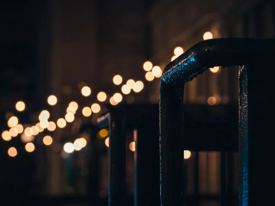 Close-up of a metal railing in focus at night, with blurred string lights glowing in the background—an inviting real estate detail highlighting modern outdoor ambiance.