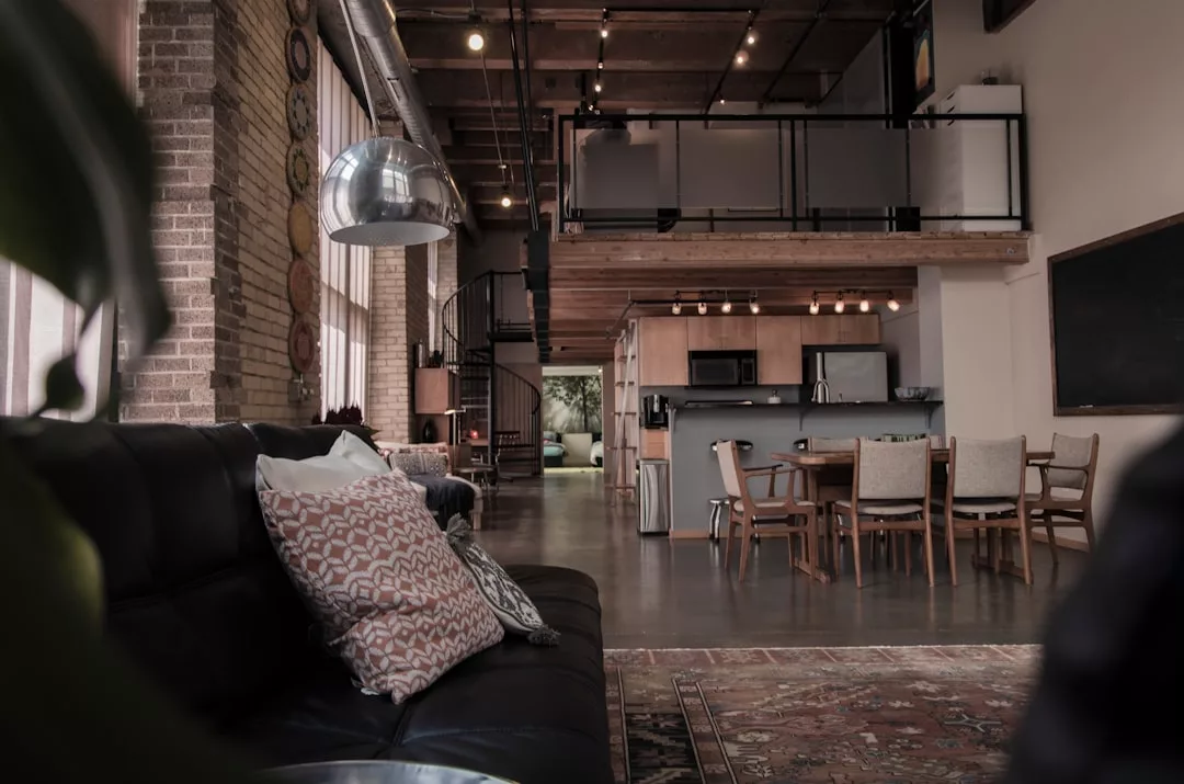 This modern real estate loft apartment features exposed brick walls, an open kitchen and dining area, a spiral staircase, and a seating area with patterned pillows.