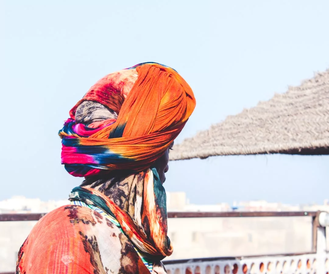 A person wearing a colorful headscarf and patterned clothing stands outdoors, facing away, with a straw shade and cityscape in the background—capturing the vibrant spirit often found in urban real estate settings.