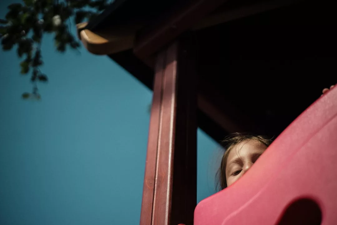 A child partially hidden behind a red playground slide looks upwards, with part of a play structure and blue sky visible in the background—a charming scene reflecting the family-friendly appeal of this real estate location.