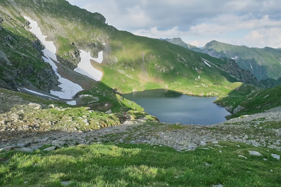 A small mountain lake surrounded by green hills, patches of snow, and rocky terrain under a partly cloudy sky—an untouched gem perfect for exclusive real estate opportunities.