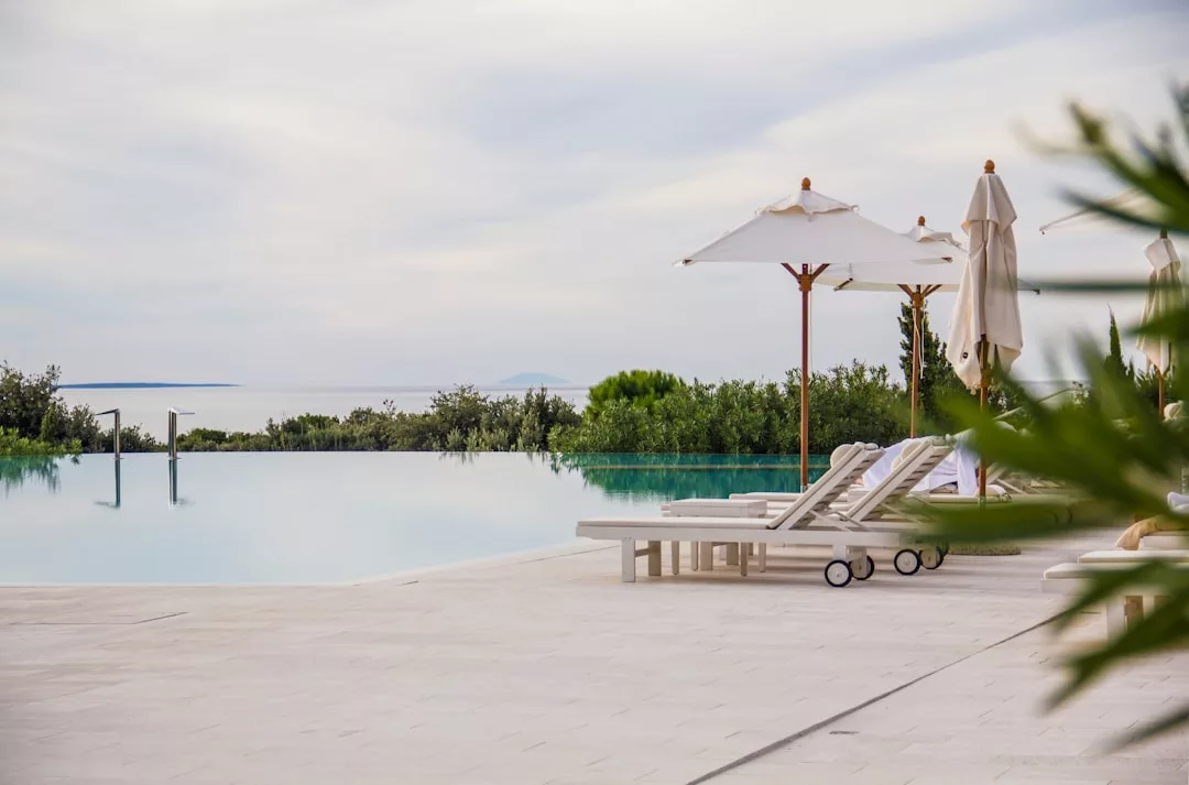 Infinity pool with sun loungers and umbrellas on a tiled deck, overlooking greenery and a calm sea—an ideal real estate opportunity for those seeking tranquility under a cloudy sky.