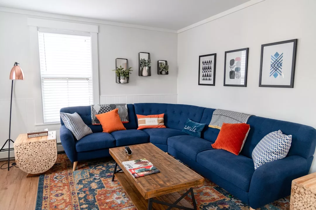 A stylish living room perfect for real estate showcases, featuring a blue sectional sofa with orange and patterned pillows, a wooden coffee table, wall art, houseplants, and a lamp beside a window with blinds.
