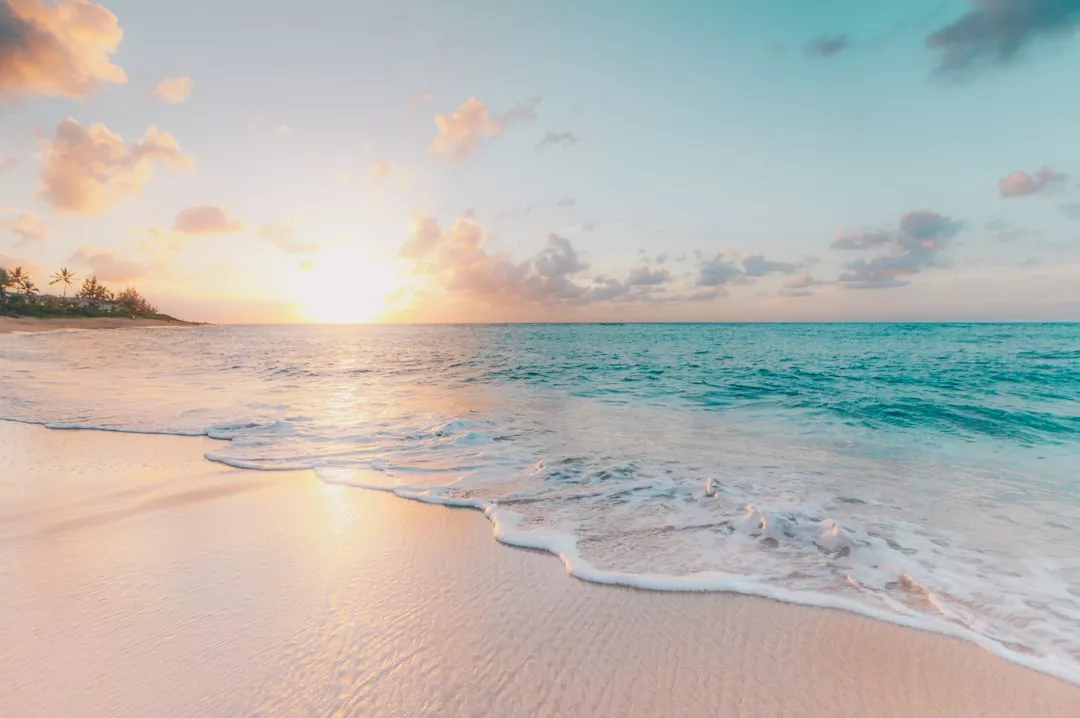 A calm beach scene at sunrise or sunset with gentle waves, soft sand, and a partly cloudy sky. Perfect for real estate dreams, the sun is low on the horizon, casting warm light over the ocean.