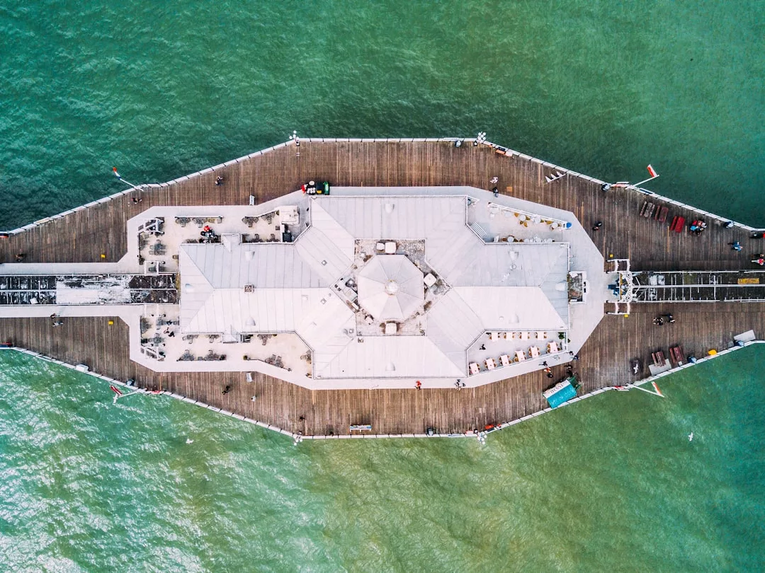 Aerial view of a large pier with a central building extending into green-blue water, people walking along the wooden boardwalk—an inviting scene that highlights unique real estate opportunities by the waterfront.