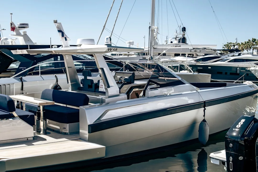 A modern silver motor yacht with a dark blue interior is docked at a marina, surrounded by other boats under clear skies—the perfect luxury feature for waterfront real estate.