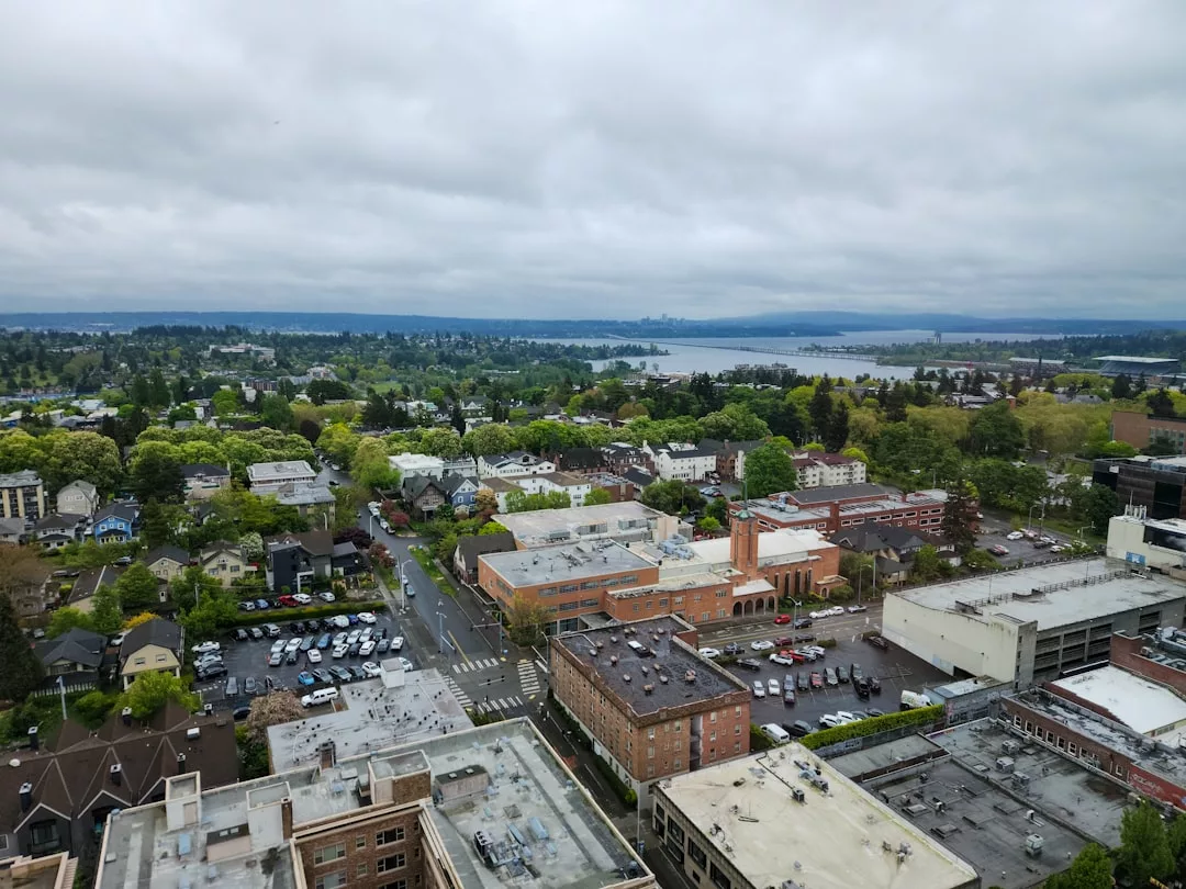 Aerial view of a suburban area with trees, buildings, parking lots, and a distant body of water under a cloudy sky—an ideal scene for real estate opportunities.