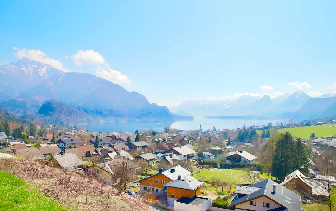A residential village with houses, green fields, and trees in the foreground, a lake in the midground, and snow-capped mountains beyond—a stunning setting perfect for real estate opportunities under a clear blue sky.