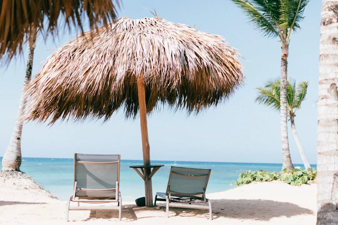 Two lounge chairs sit under a large thatched umbrella on a sandy beach, with palm trees and the ocean in the background—a true paradise setting perfect for luxurious real estate retreats.