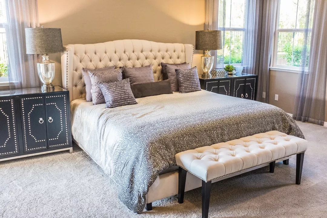 A neatly made bed with a tufted headboard, gray and beige bedding, decorative pillows, two side tables with lamps, a bench at the foot, and windows with sheer curtains create an inviting real estate showcase.