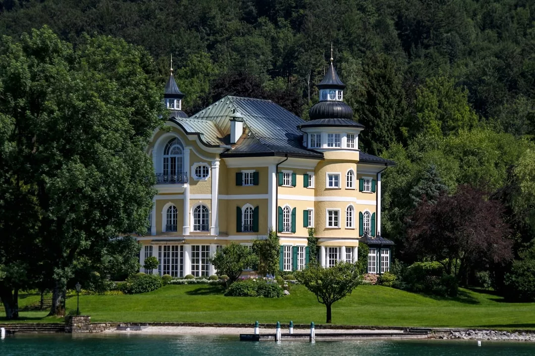 A stunning piece of real estate, this large yellow three-story house with multiple turrets and a green roof sits by the water, surrounded by lush trees and manicured lawns.