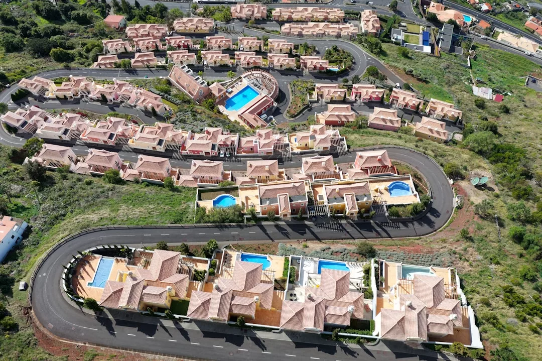 Aerial view of a residential neighborhood with rows of houses, several with private pools, and a shared central pool—an inviting real estate haven surrounded by curving roads and lush greenery.