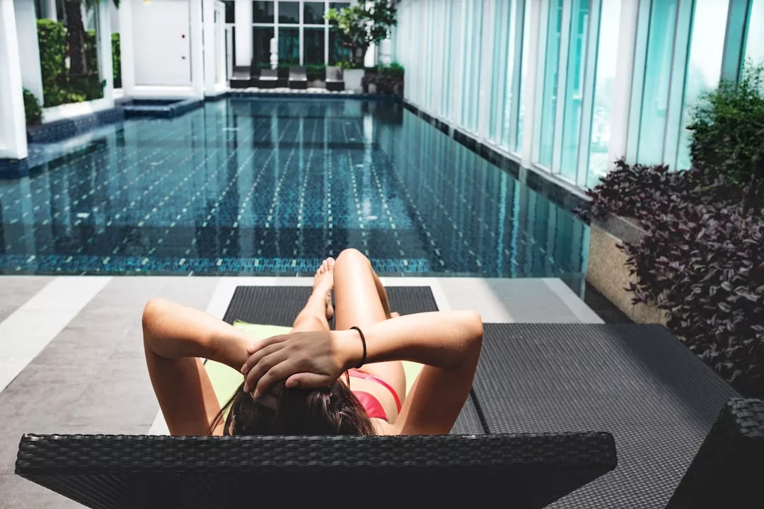 A person in swimwear reclines on a lounge chair beside an indoor swimming pool, with glass walls and plants offering a glimpse of upscale real estate ambiance in the background.