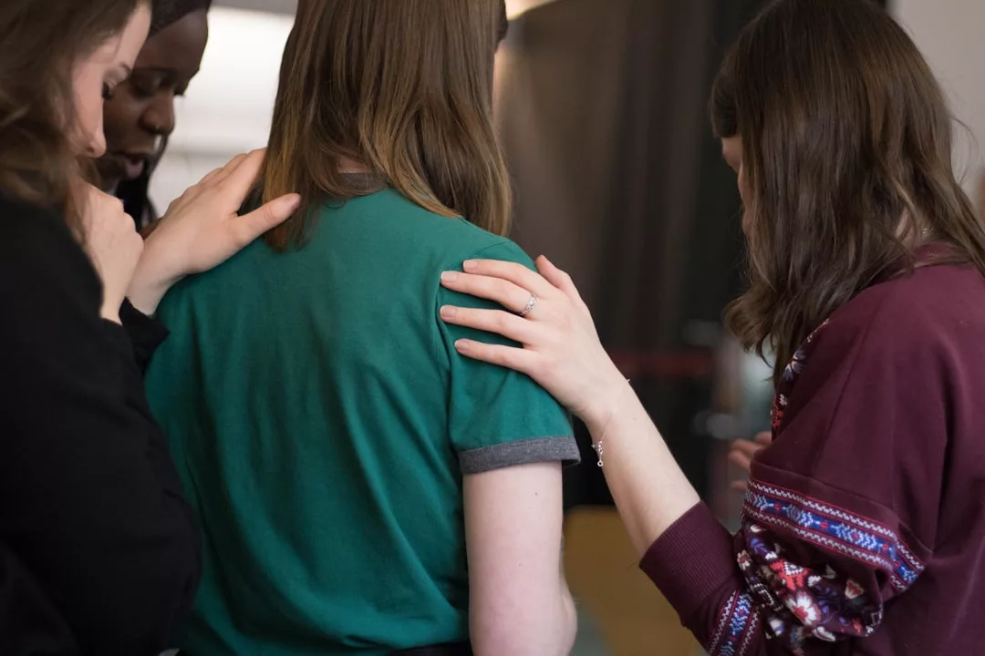Four people stand close together, with three placing their hands supportively on the back and shoulder of the person in the green shirt, suggesting comfort or reassurance—much like a real estate team guiding a client through important decisions.