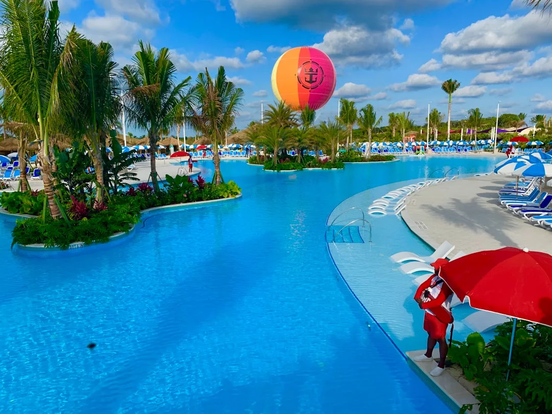 A large pool with palm trees, lounge chairs, and umbrellas creates a relaxing oasis. A lifeguard stands under a red umbrella, while a colorful hot air balloon with a real estate logo floats in the background.