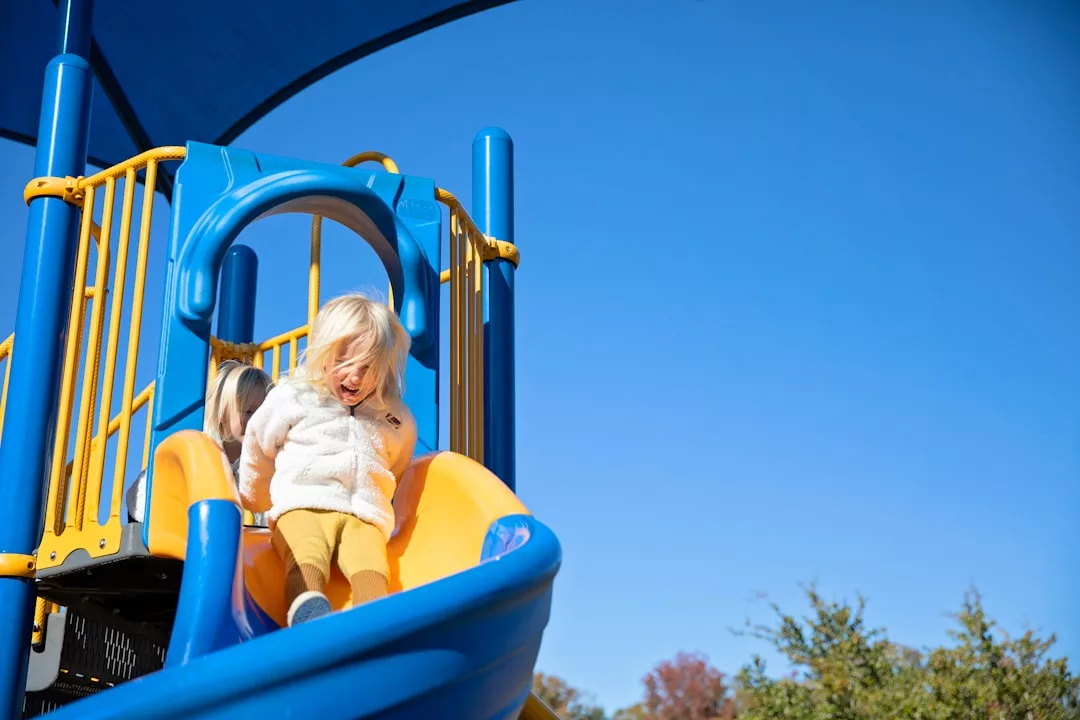Two young children slide down a yellow and blue playground slide under a clear blue sky, highlighting the family-friendly amenities often sought after in real estate.