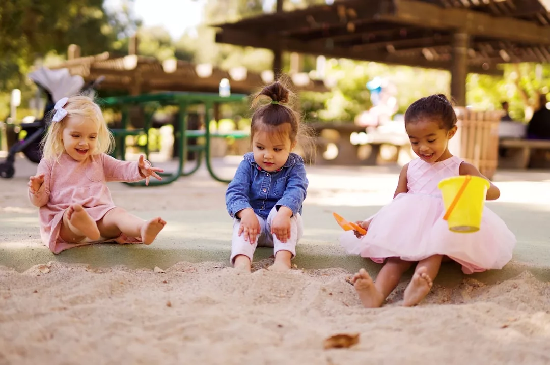 Three young children sit at the edge of a sandbox outdoors, enjoying playtime near their family’s real estate. Two wear pink dresses, one a denim jacket, as they dig in the sand with a yellow bucket.