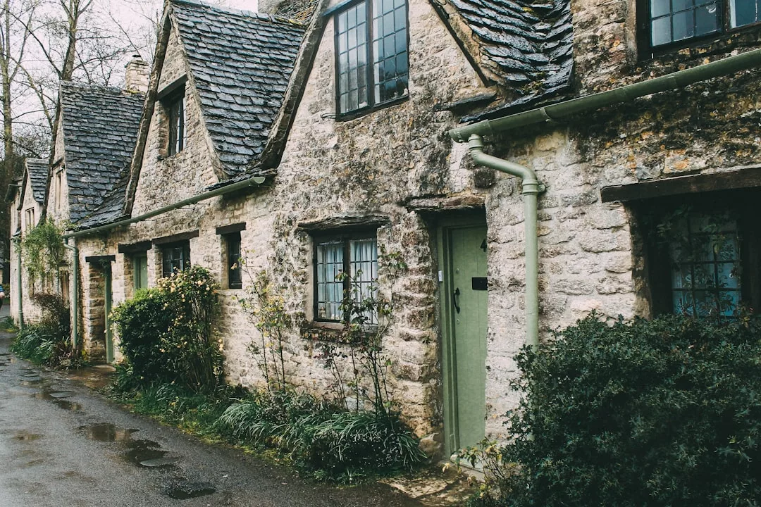 Row of old stone cottages with slate roofs and small windows, bordered by greenery, line a quiet, wet street—ideal real estate for those seeking charm and tranquility.