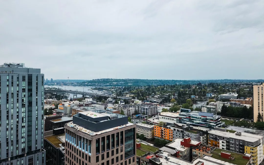 Aerial view of a cityscape featuring mid-rise buildings, roads, and greenery under a cloudy sky; perfect for real estate opportunities with a body of water and distant hills visible in the background.