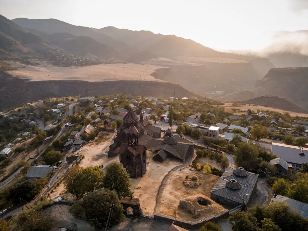 Aerial view of a historic church complex, ideal for real estate opportunities, surrounded by small houses and trees in a valley with mountains and dry fields under a hazy sky.