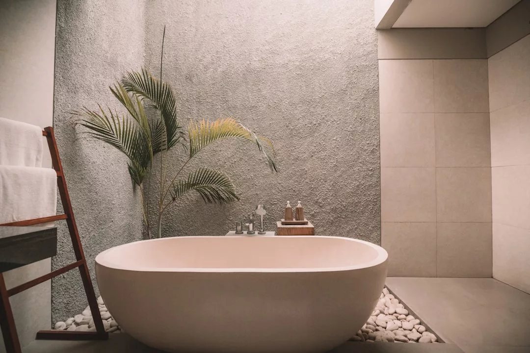 A modern bathroom perfect for real estate showcases, featuring a freestanding oval bathtub on white pebbles, a potted plant, and neatly folded towels on a wooden rack against textured walls.