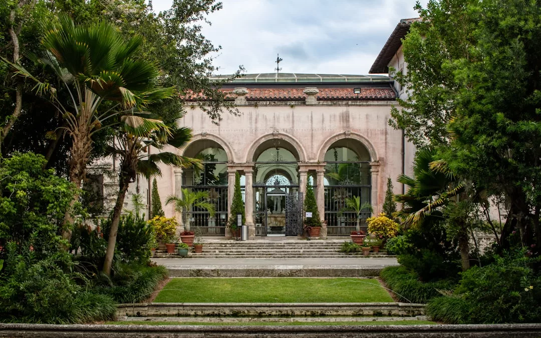 A grand historic building with three large arched doorways, surrounded by lush trees, potted plants, and manicured lawns—an exceptional real estate opportunity blending timeless elegance with natural beauty.