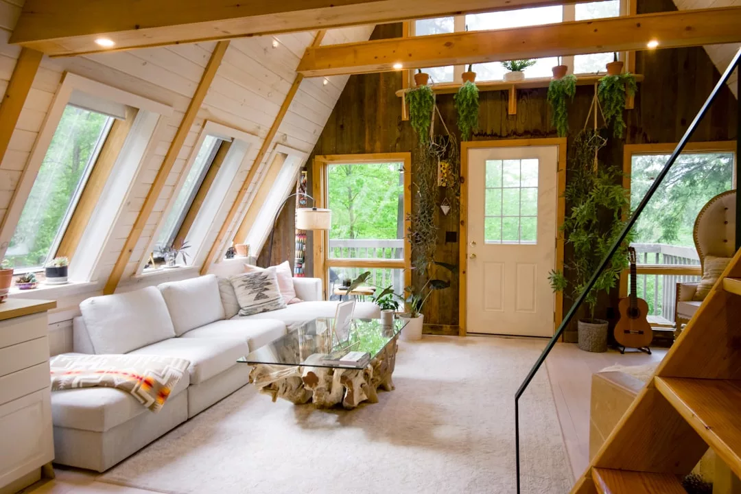 A bright A-frame living room perfect for real estate listings, featuring large windows, a white sectional sofa, glass coffee table, hanging plants, and a staircase leading upstairs.