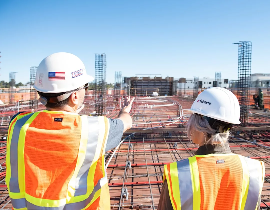 Two construction workers in safety vests and hard hats stand on a real estate construction site, with one gesturing toward the building framework ahead.