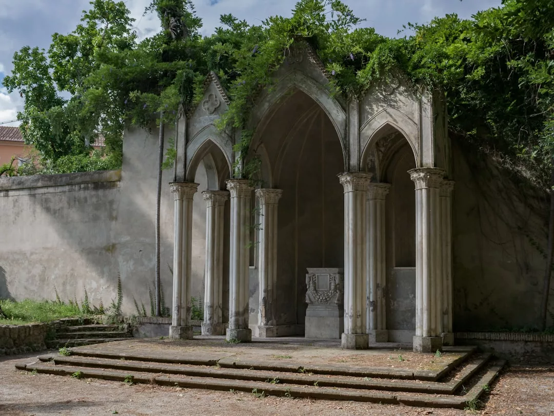 A small stone stage with three gothic arches and columns, partially covered by greenery, stands in front of a weathered wall and trees—a charming feature for unique real estate settings.