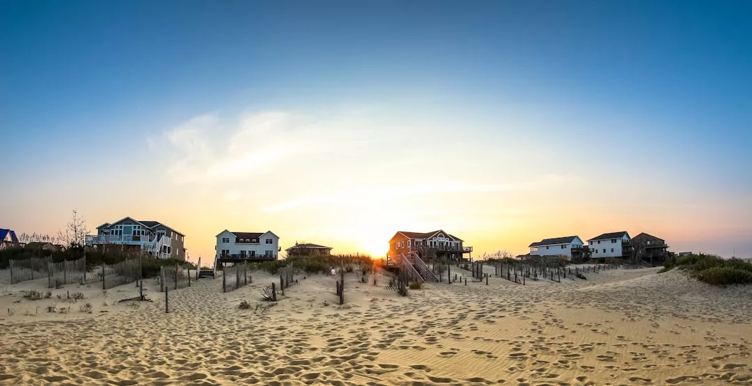Five beach houses are lined up behind sand dunes and a fence, their silhouettes glowing in the sunset—a picturesque scene perfect for real estate enthusiasts, with a clear blue sky stretching above.