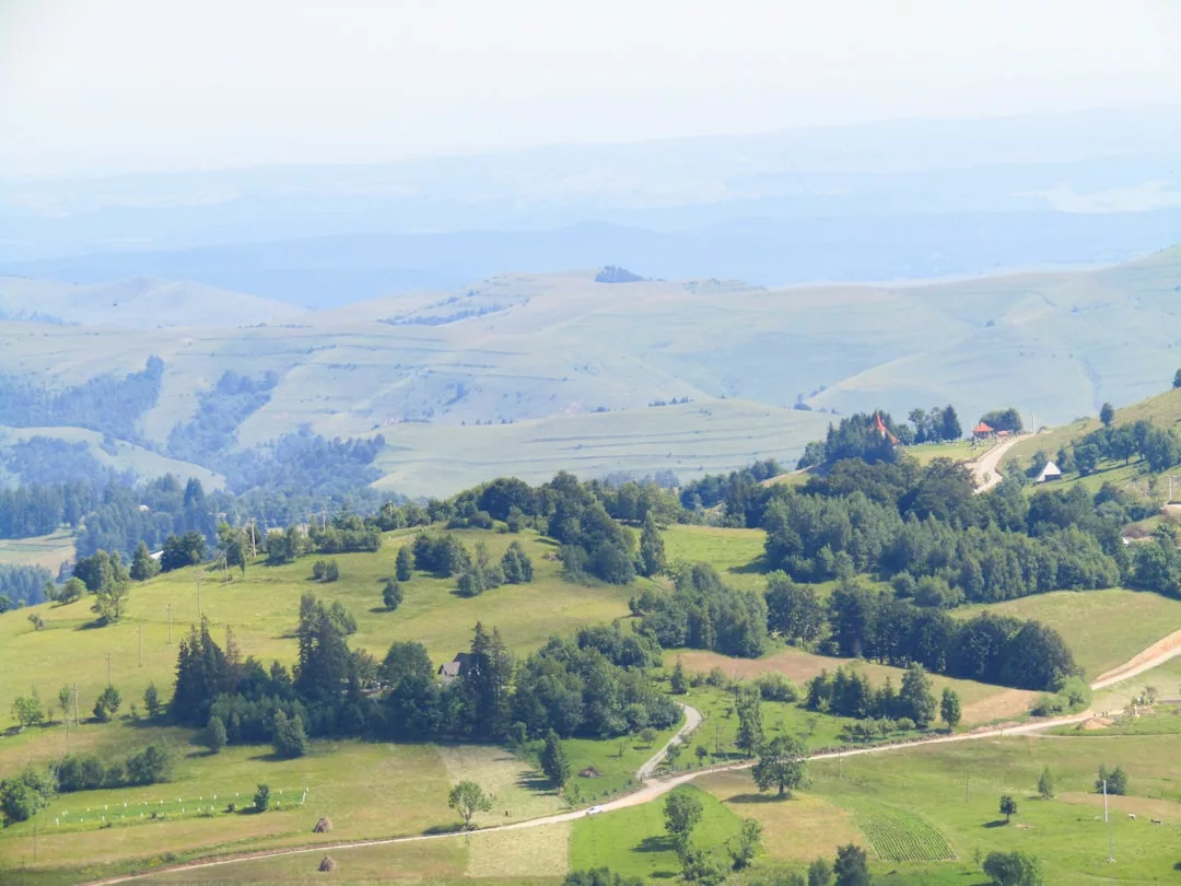 Aerial view of rolling green hills dotted with trees and houses, winding dirt roads, and distant blue-tinted mountains under a hazy sky—an idyllic real estate opportunity in a picturesque landscape.