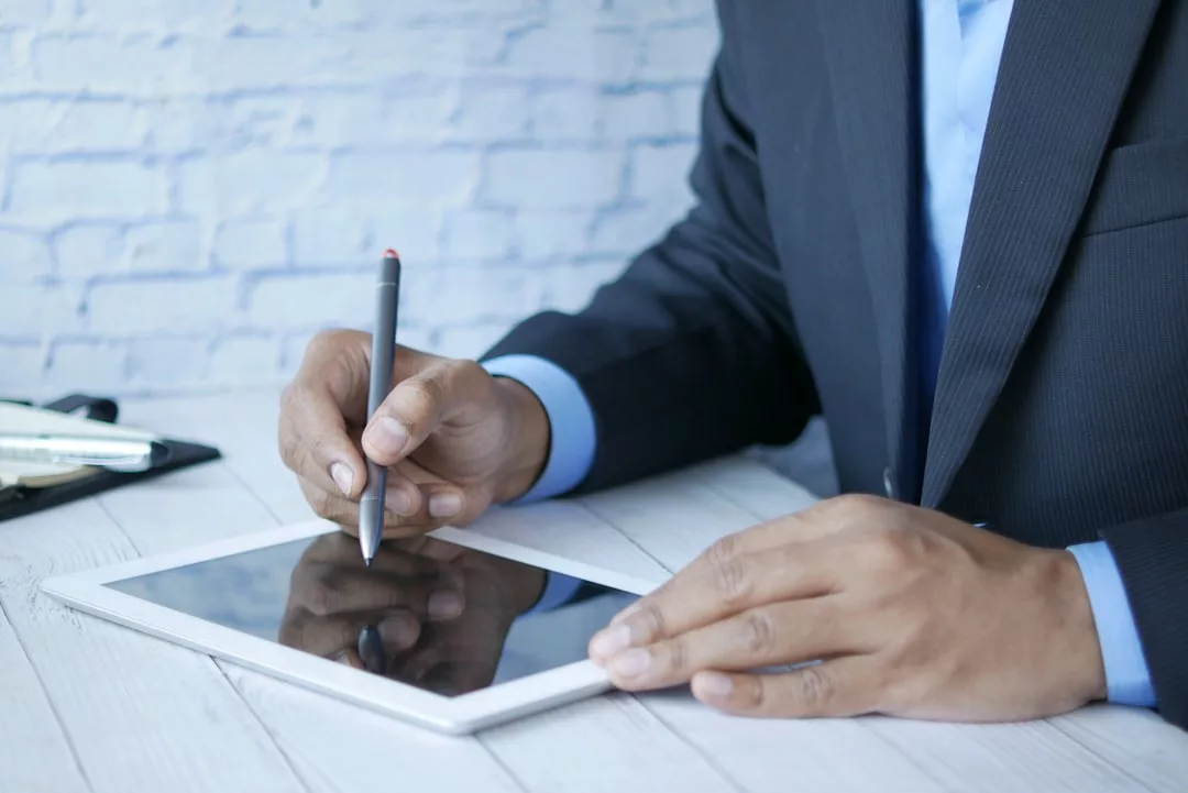 A person in a suit uses a stylus to write on a tablet placed on a white wooden table, preparing real estate documents.