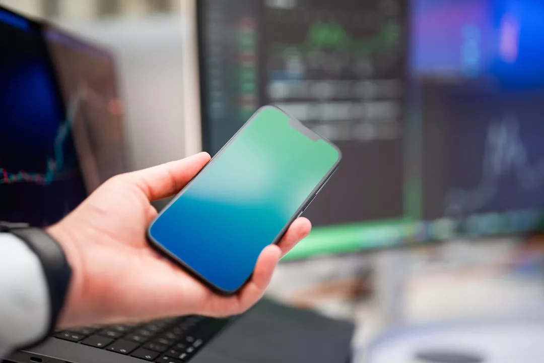 A person holds a smartphone with a blank screen in front of computer monitors displaying financial charts and real estate market data.