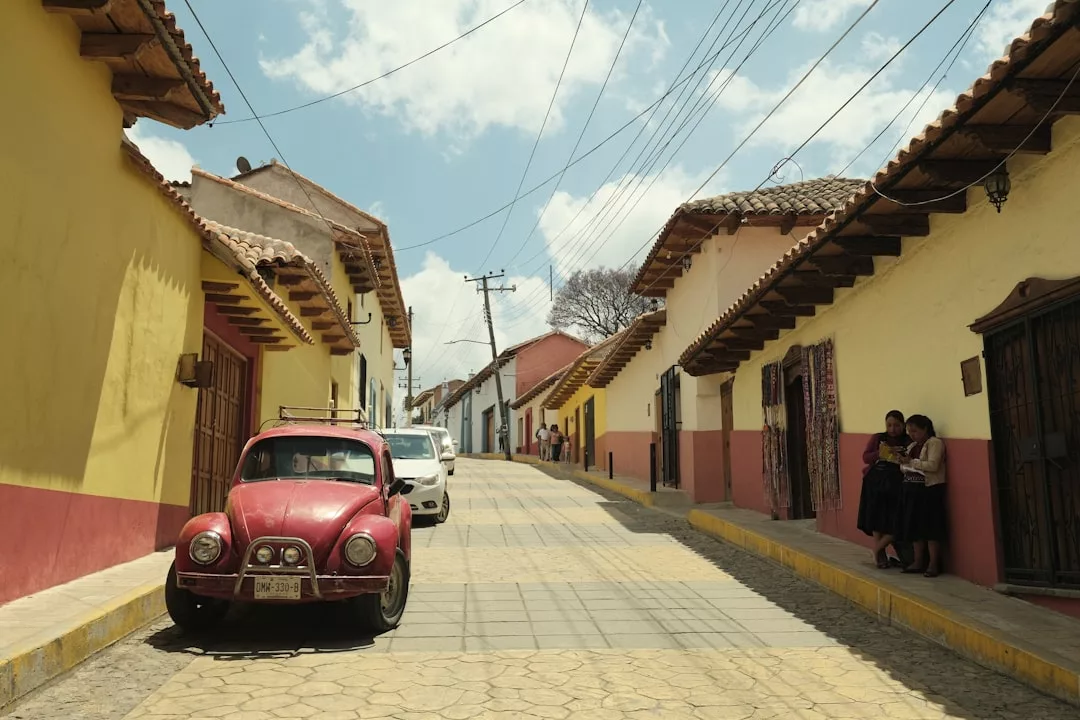 A red Volkswagen Beetle is parked on a cobblestone street lined with colorful buildings; three people stand talking near a doorway on the right, possibly discussing local real estate opportunities.